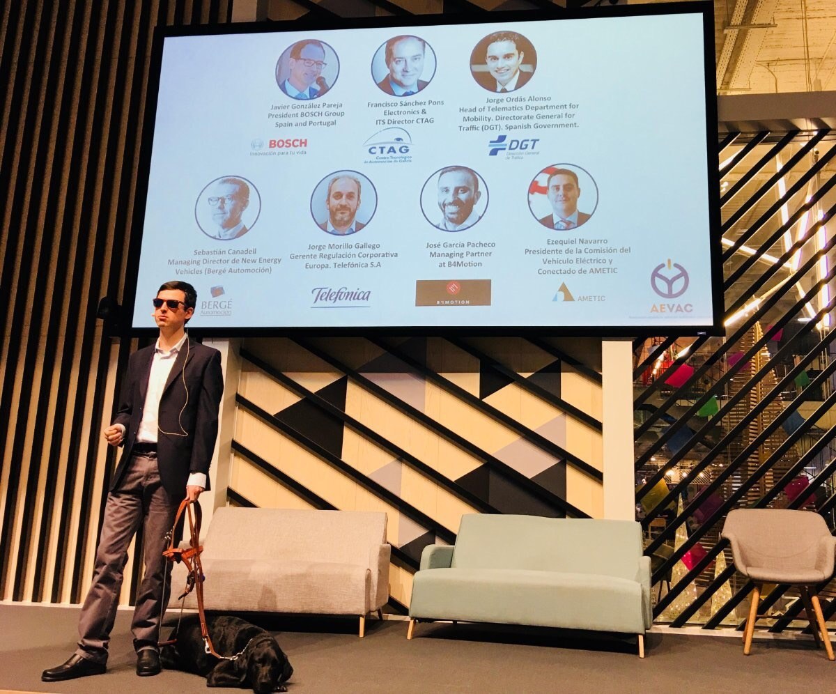 Aitor Fernández con su perro guía en el escenario de Campus Madrid; la pantalla muestra a los participantes del encuentro de presentación de AEVAC.