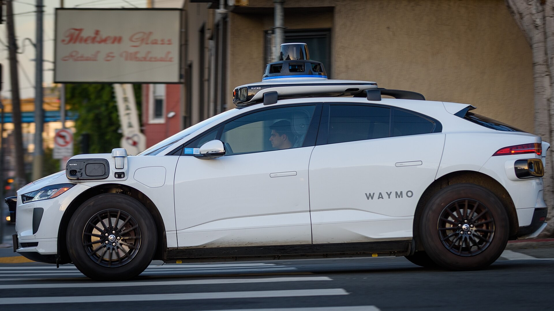 A Waymo autonomous car, a white Jaguar I-Pace with sensors on the roof, driving on a street.