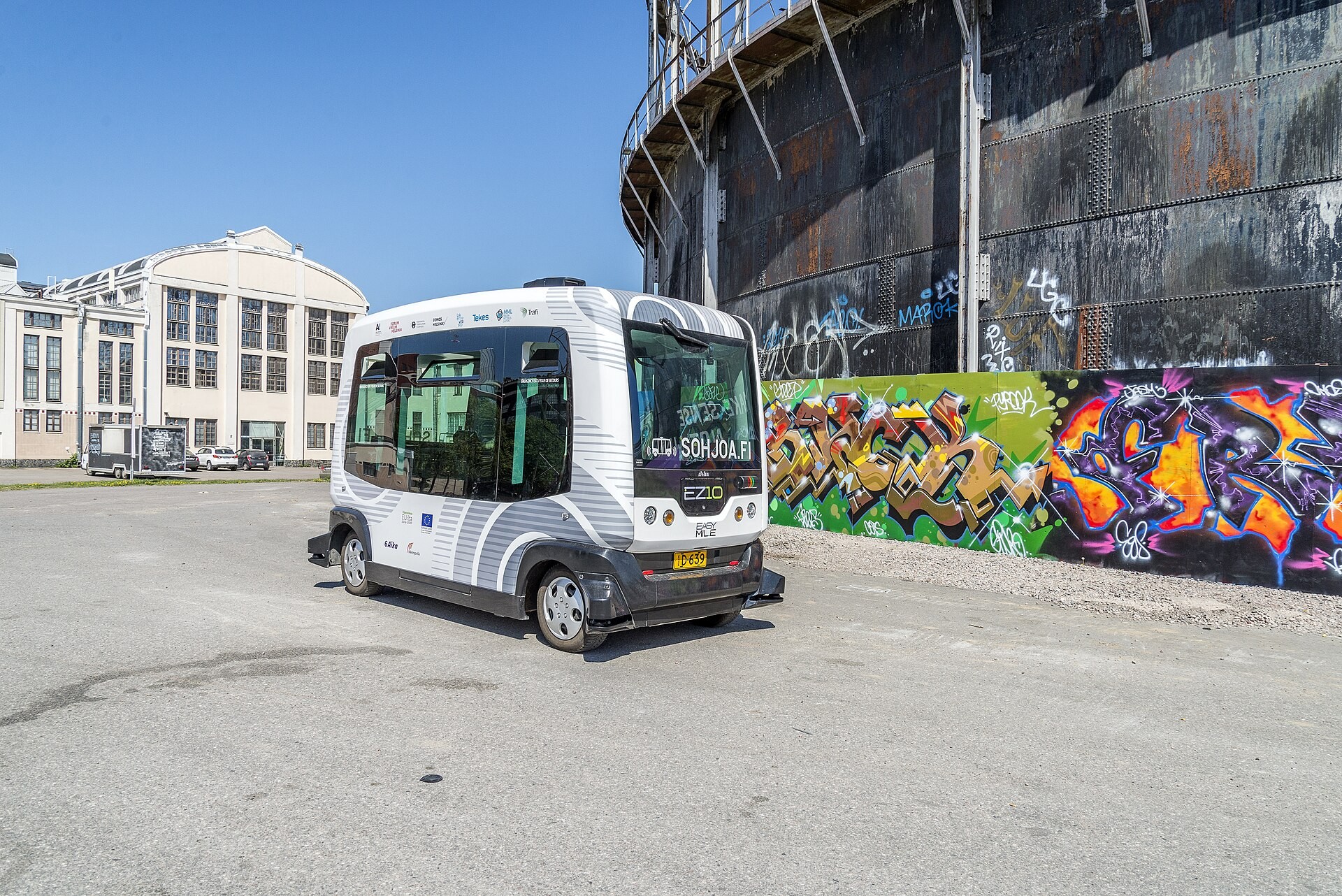 Pequeño autobús autónomo EZ10 blanco y gris, el mismo modelo que usó ALSA en Cantoblanco, parado junto a una nave industrial en Helsinki.